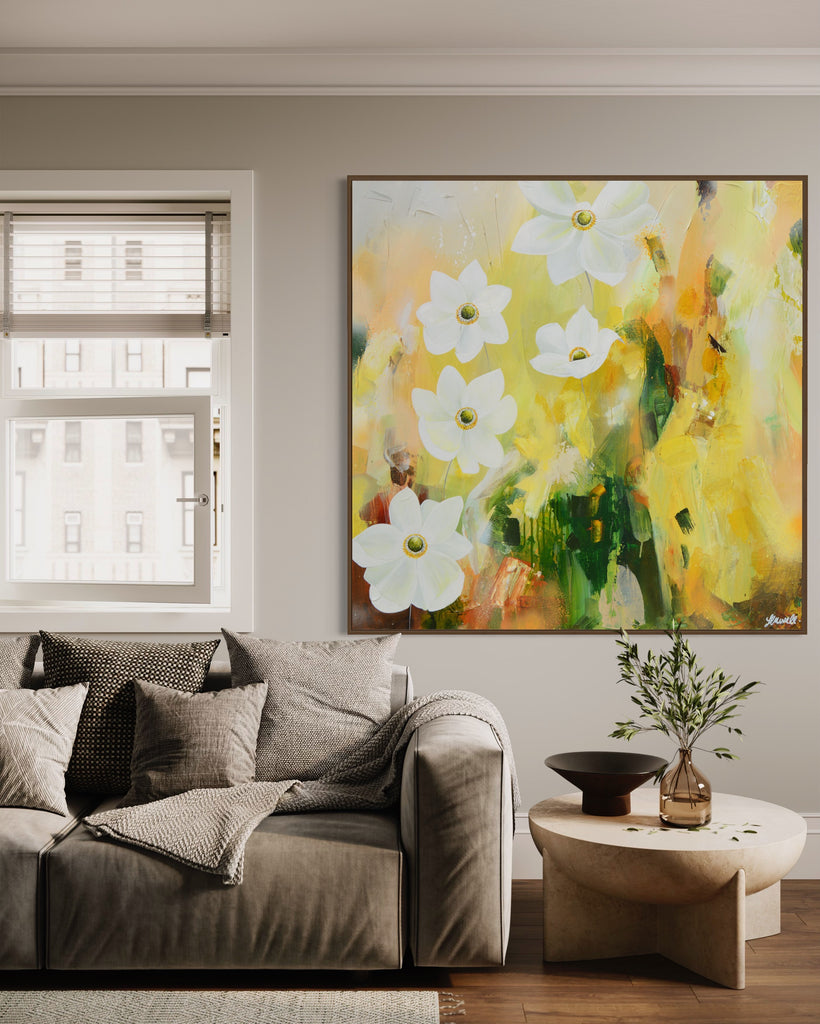 A 'Fresh Start' original acrylic painting by Jasmine Marshall framed in black/oak. Canvas painting features white anemone flowers atop cream, lime green, yellow, and brown abstract details, featured in a neutral living room with a grey sofa and cushions and a wooden coffee table.