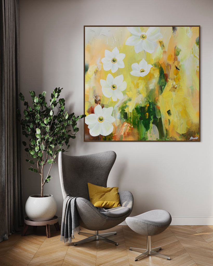 A 'Fresh Start' original acrylic painting by Jasmine Marshall framed in black/oak. Canvas painting features white anemone flowers atop cream, lime green, yellow, and brown abstract details, featured in a neutral home interior with a grey chair, yellow cushion, grey curtains and a plant..