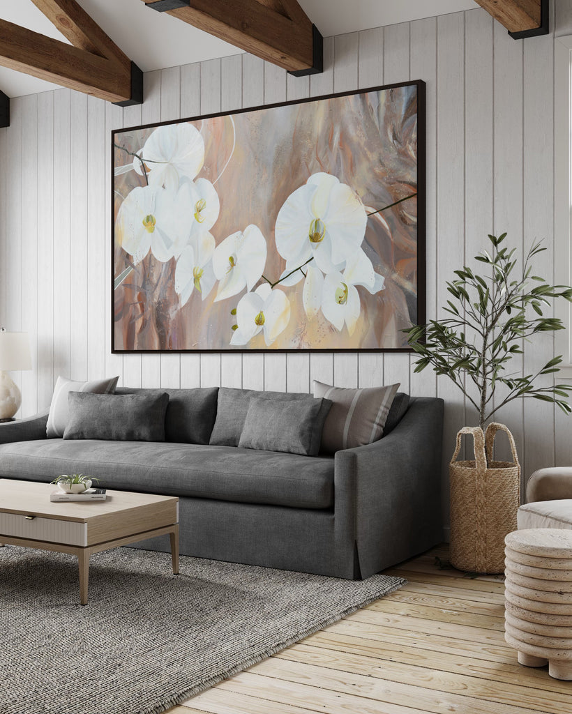 'Natural Elegance' original painting on a cotton canvas by Jasmine Marshall, framed in black/oak. Features white orchids on a neutral brown, beige abstract background. Displayed in a living room with wooden ceiling beams, grey sofa, plant and wooden coffee table.