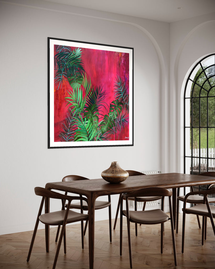 ‘The Foliage' Fine Art Print by Jasmine Marshall, framed in black/oak. Premium Giclée print features tropical leaves over an abstract pink, red and orange painting. Displayed in a neutral dining room with a wooden dining table and chairs and big glass doors.