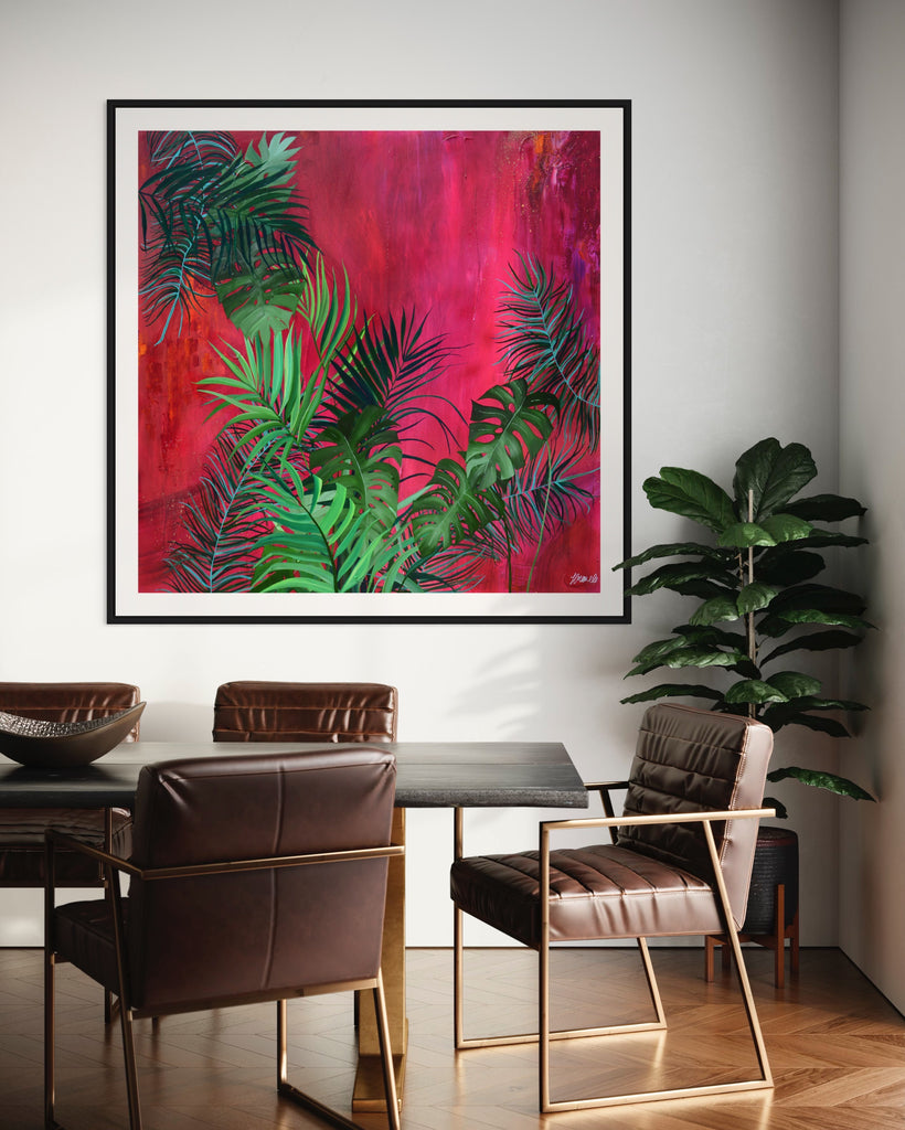 ‘The Foliage' Fine Art Print by Jasmine Marshall, framed in black/oak. Premium Giclée print features tropical leaves over an abstract pink, red and orange painting. Displayed in a kitchen with brown wooden dining table and plant.