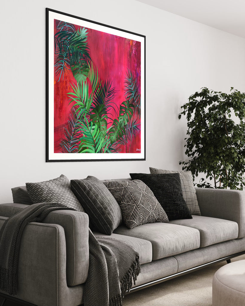 ‘The Foliage' Fine Art Print by Jasmine Marshall, framed in black/oak. Premium Giclée print features tropical leaves over an abstract pink, red and orange painting. Displayed in a living room with grey sofas and blanket and plant.