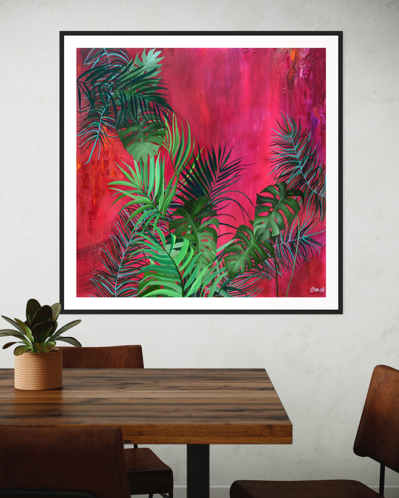 ‘The Foliage' Fine Art Print by Jasmine Marshall, framed in black/oak. Premium Giclée print features tropical leaves over an abstract pink, red and orange painting. Displayed in a kitchen with wooden dining table and plant on.