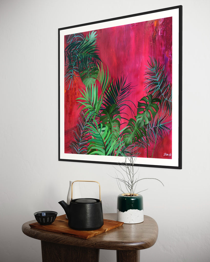 ‘The Foliage' Fine Art Print by Jasmine Marshall, framed in black/oak. Premium Giclée print features tropical leaves over an abstract pink, red and orange painting. Displayed in a neutral home interior.