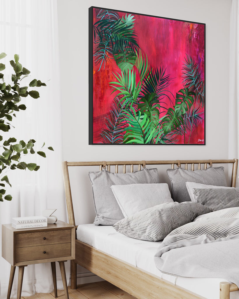 'The Foliage' original painting on a cotton canvas by Jasmine Marshall, framed in black/oak. Features tropical leaves over an abstract pink, red and orange painting Displayed in a neutral home interior above a wooden bed frame.