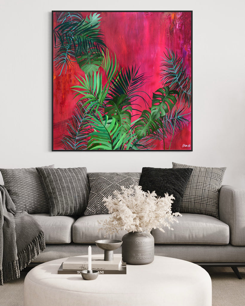 'The Foliage' original painting on a cotton canvas by Jasmine Marshall, framed in black/oak. Features tropical leaves over an abstract pink, red and orange painting Displayed in a living room with grey sofa, cushions and blanket and white coffee table.