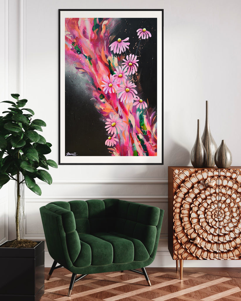 ‘The Laughter of Daisies' Fine Art Print by Jasmine Marshall, framed in black/oak. Premium Giclée print features pink daises on a colourful abstract details on a black background. Displayed next to a green velvet arm chair plant and bronze side table.