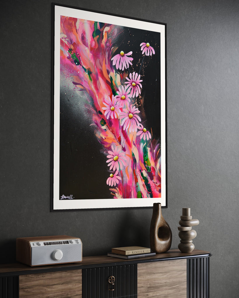 ‘The Laughter of Daisies' Fine Art Print by Jasmine Marshall, framed in black/oak. Premium Giclée print features pink daises on a colourful abstract details on a black background. Displayed on a dark grey wall with wooden unit.