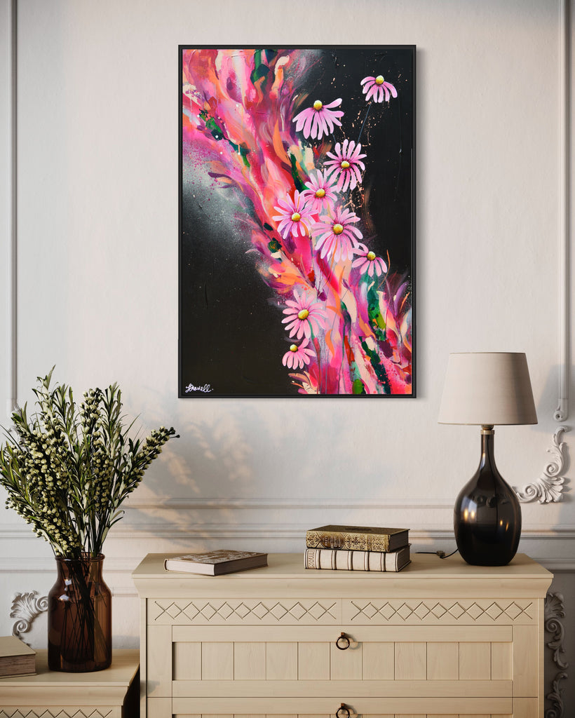 'The Laughter of Daisies' original painting on a cotton canvas by Jasmine Marshall, framed in black/oak. Features pink daises on a colourful abstract details on a black background. Displayed in a neutral home interior above a chest of drawers with books and plants on.