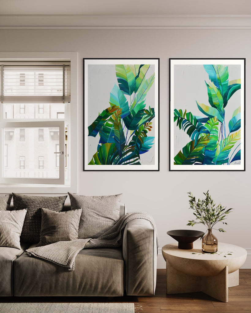 ‘Tropic Duo' Fine Art Print by Jasmine Marshall, framed in black/oak. Premium Giclée print features tropical blue and green leaves on a grey background. Displayed in a living room with grey sofa and wooden side table with plant.