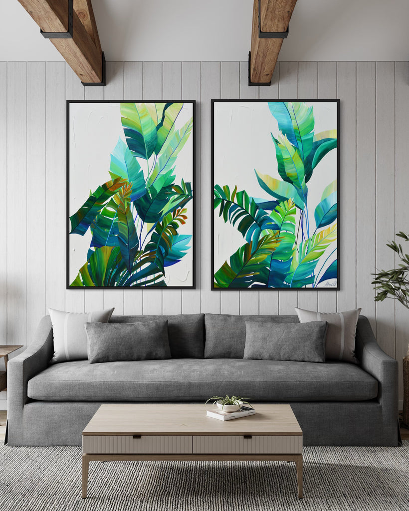 'Tropic Duo' original painting on a cotton canvas by Jasmine Marshall, framed in black/oak. Features tropical blue and green leaves on a grey background. Displayed in a neutral home interior with grey sofa, wooden ceiling beams, grey cushions and a green plant.