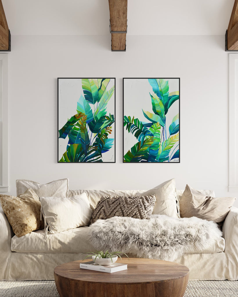 'Tropic Duo' original painting on a cotton canvas by Jasmine Marshall, framed in black/oak. Features tropical blue and green leaves on a grey background. Displayed in a neutral living room with wooden ceiling beams and a wooden coffee table with a book and plant on, white sofa and cushions.