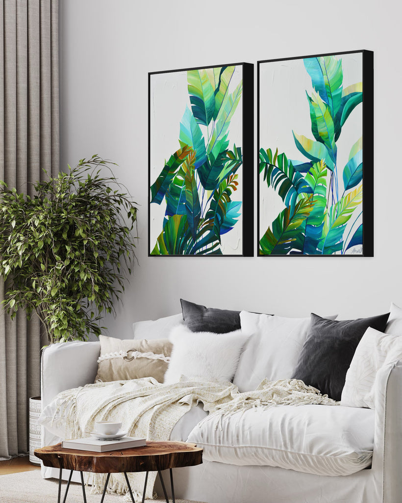 'Tropic Duo' original painting on a cotton canvas by Jasmine Marshall, framed in black/oak. Features tropical blue and green leaves on a grey background. Displayed in a neutral home interior with white sofa, grey cushions, grey curtains, plant, wooden coffee table.