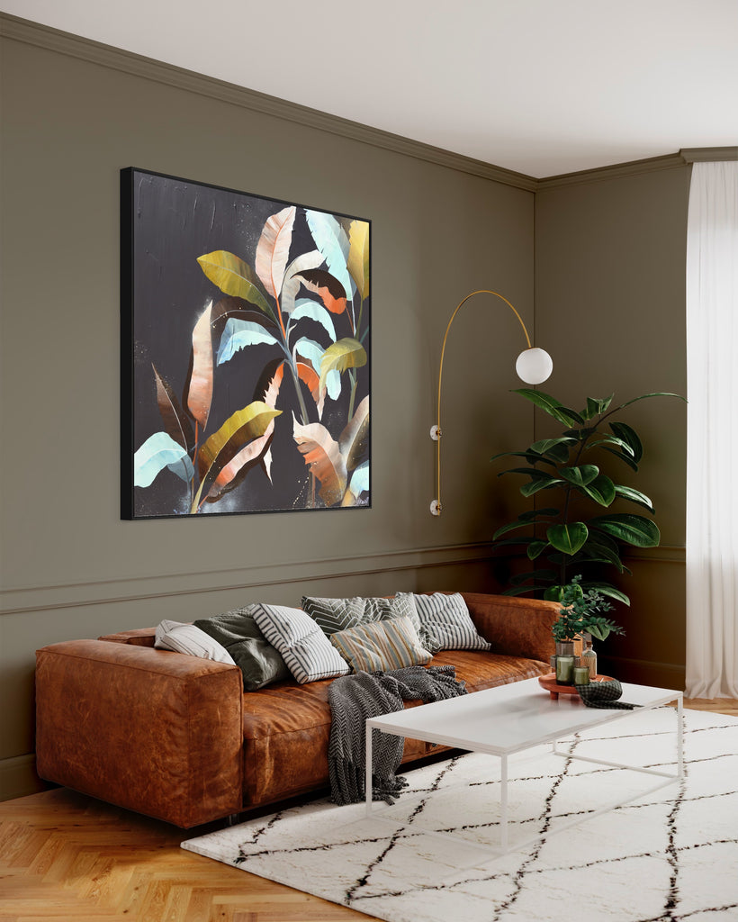 'Under the Canopy' original painting on a cotton canvas by Jasmine Marshall, framed in black/oak. Features a palm tree on a brown background with abstract details. Displayed on a green wall in a living room with a brown sofa, white cushions, white rug and green plant.